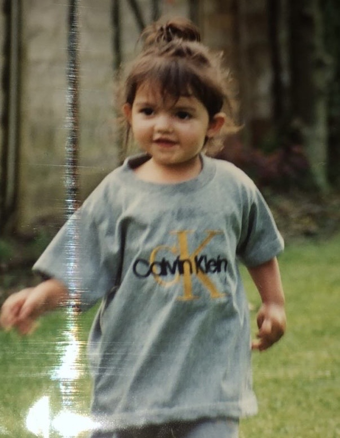 Ellie as a toddler, mid-stride on a garden lawn, hair in a topknot, wearing an enormous Calvin Klein t-shirt.