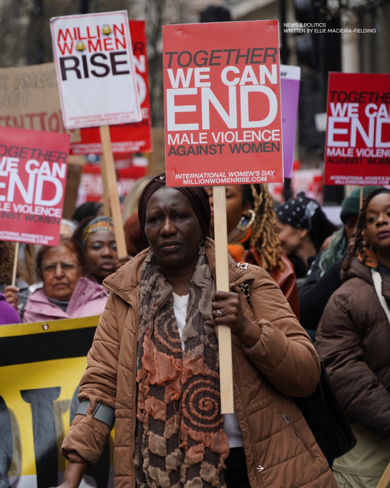 A woman in a headwrap and brown coat carries a red 'Together we can end male violence against women' placard through the march.