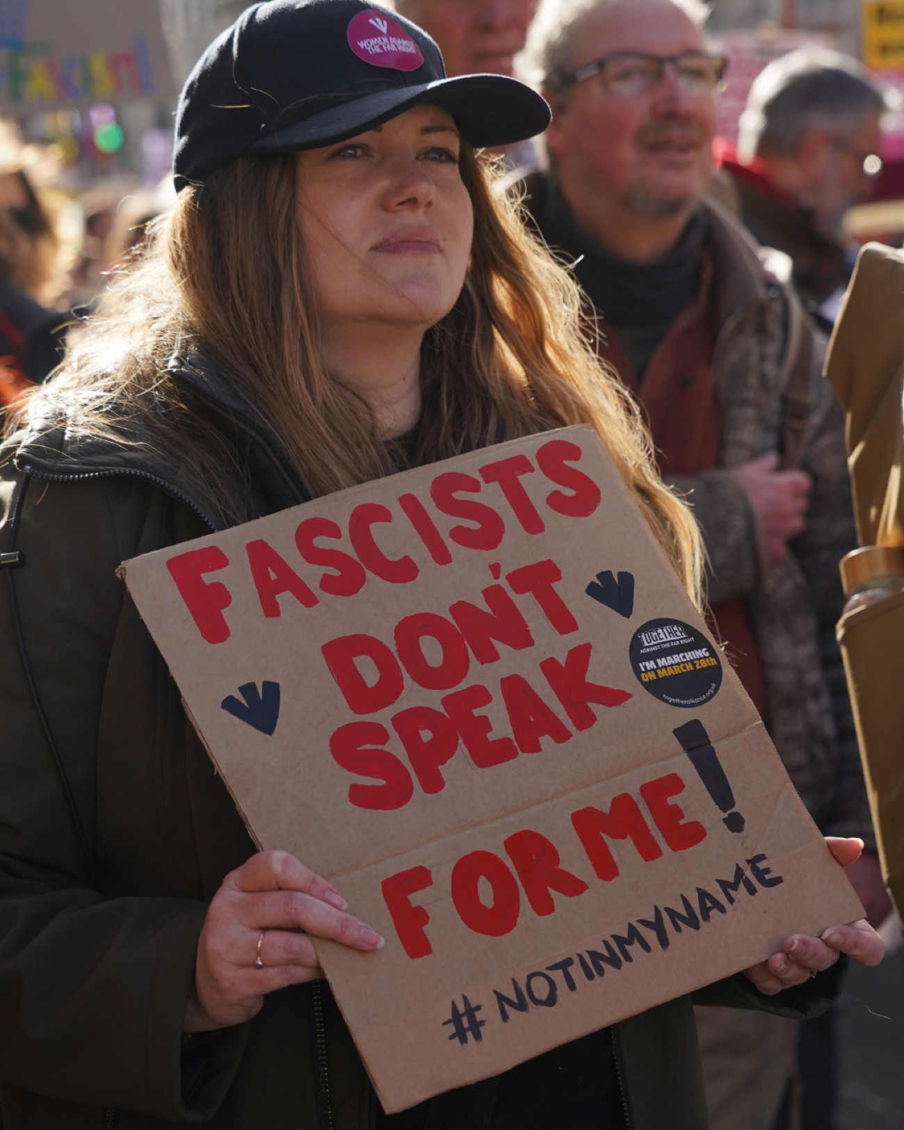 A woman in a baseball cap holds a hand-painted cardboard sign reading 'Fascists don't speak for me. #NotInMyName'.