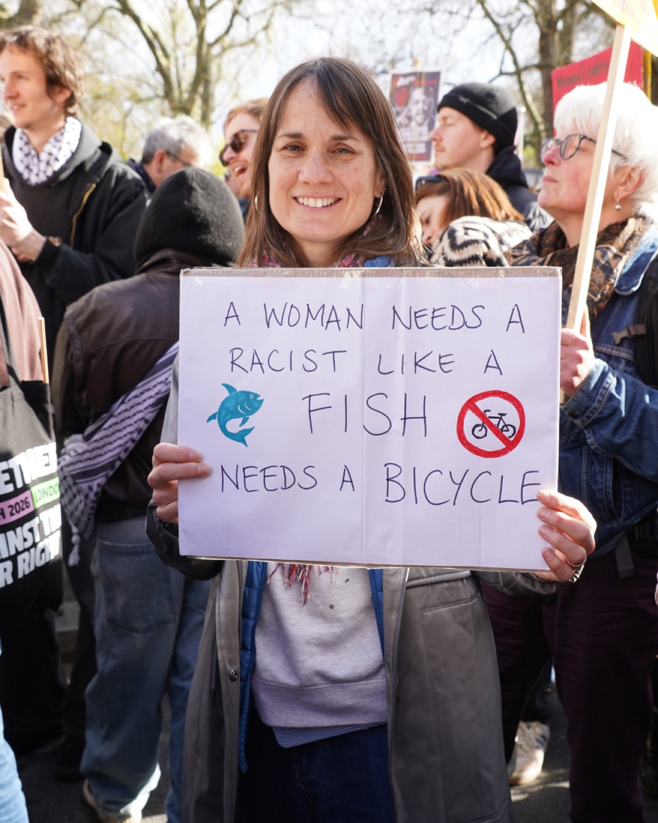 A smiling protester holds a sign reading 'A woman needs a racist like a fish needs a bicycle'.
