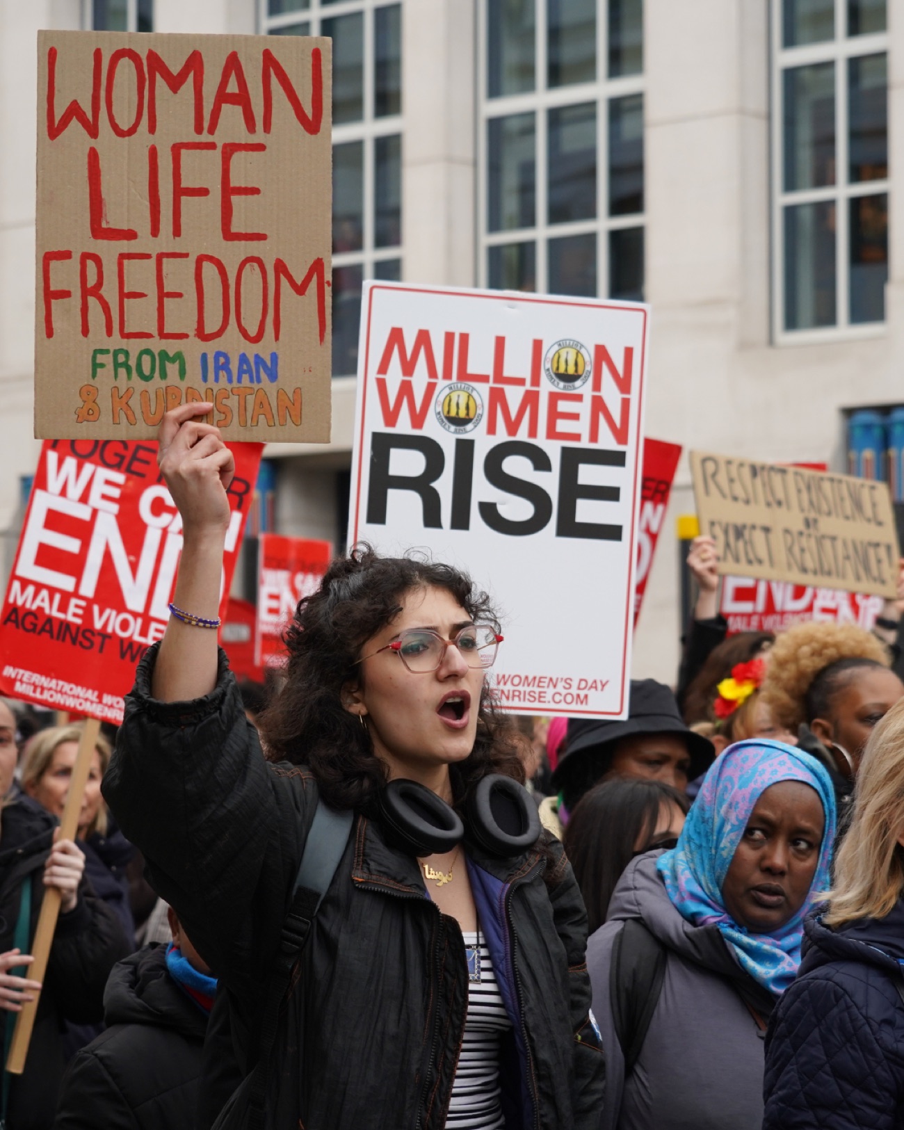 A young woman shouts while raising a cardboard sign reading 'Woman Life Freedom, from Iran and Kurdistan' above the crowd.
