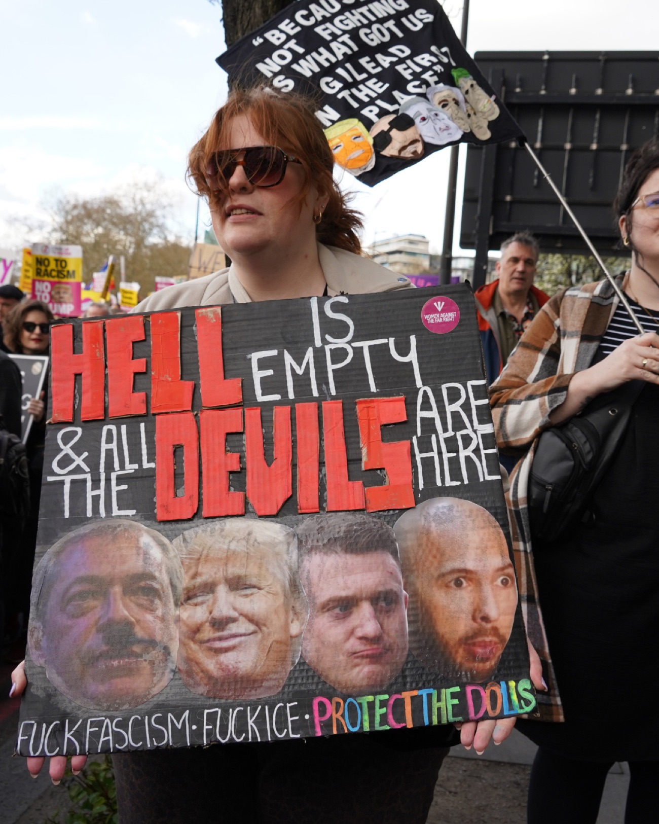 A protester holds a large hand-lettered sign reading 'Hell is empty and all the devils are here' with cut-out faces along the bottom.