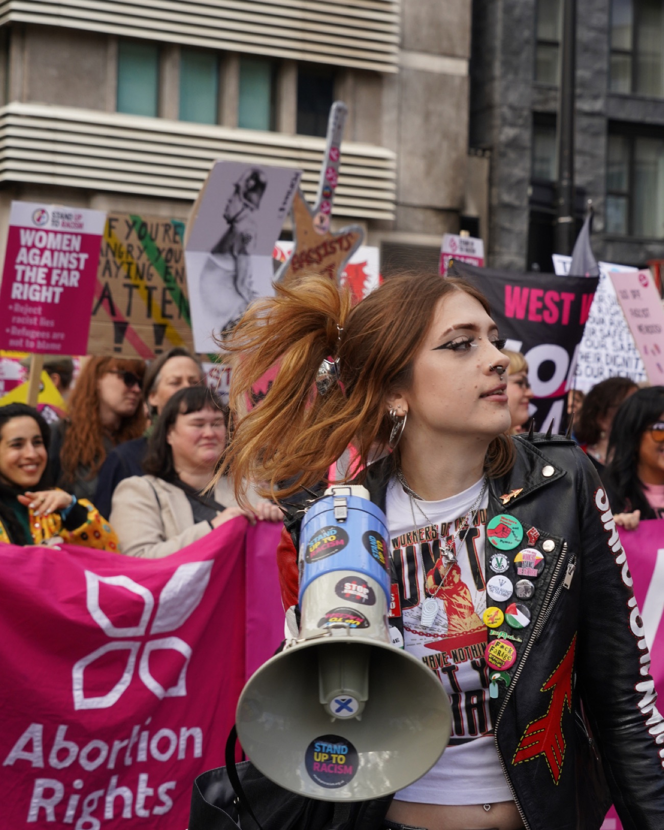 A young woman with eyeliner and a studded leather jacket carries a megaphone past a pink Abortion Rights banner.
