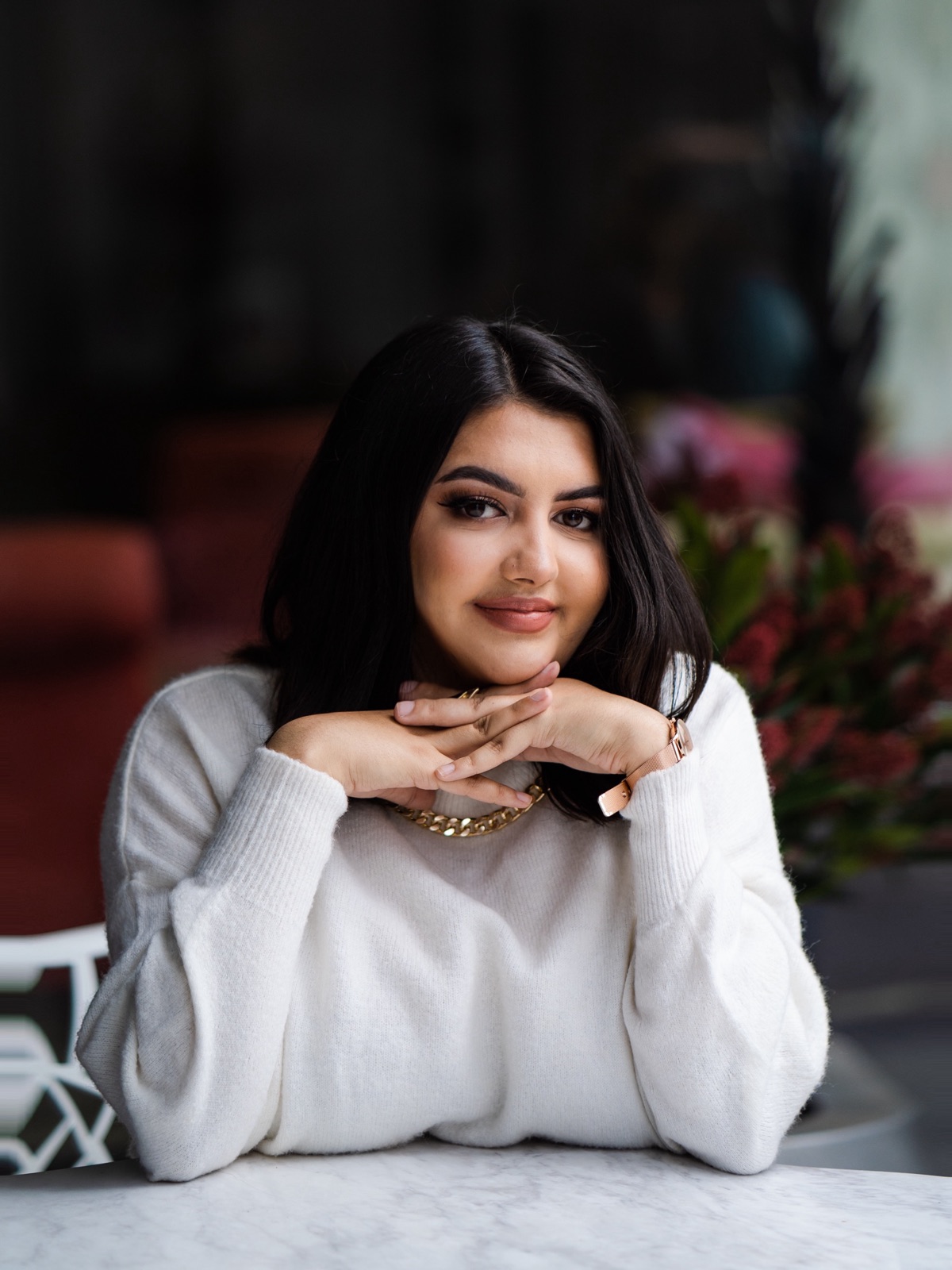 Portrait of Ellie resting her chin on her hands at a marble table, wearing a cream jumper and gold chain.